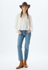 Blusa blanca texturizada con un dobladillo en peplum, frente abotonado, combinada con jeans rectos de color azul claro y botas de tobillo color marrón. Sombrero de fieltro marrón como acento.