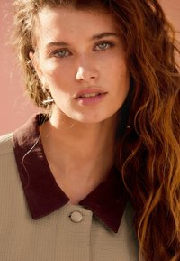 Chemise à col en beige clair texturé avec un col d'un bourgogne foncé, présentant une fermeture à boutons à l'avant et une texture capillaire visible.