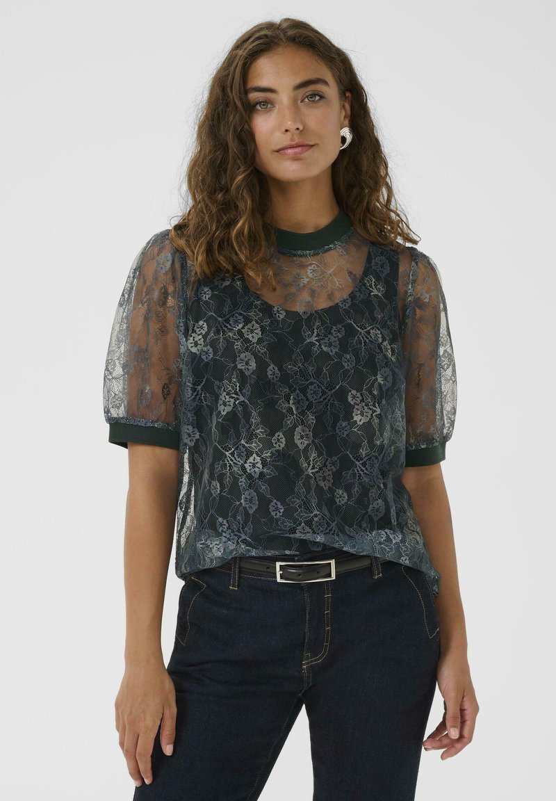 Frau mit lockigem braunem Haar, die eine durchsichtige, blumengemusterte Spitzenbluse über einem dunklen Oberteil, eine dunkelblaue Jeans, einen schwarzen Gürtel und silberne Creolen trägt.