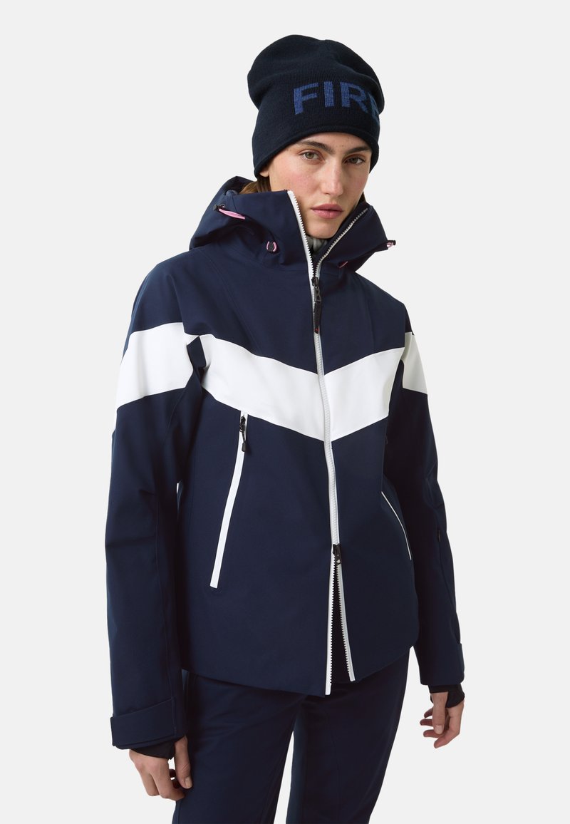 Granatowa kurtka narciarska z białym wzorem w kształcie szewrona, wyposażona w zamek błyskawiczny z przodu, kaptur i kieszenie na zamek. Do kompletu czarna czapka typu beanie.