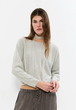 Junge Frau mit langen braunen Haaren trägt ein hellgraues langärmeliges Oberteil mit Ausschnitt am Hals und karierte Hose mit Tunnelzug, steht vor weißem Hintergrund.