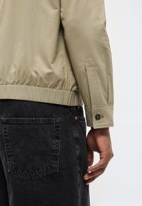 Μανίκι μπεζ σακακιού με μανσέτα με κουμπί πάνω από την τσέπη μαύρου τζιν, δείχνοντας το δεξί χέρι χαλαρό δίπλα στο ισχίο.