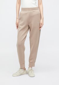 Μπεζ παντελόνια φόρμας με στενή γραμμή στα πόδια, ελαστική μέση και πλαϊνές φερμουάρ. Υφασμάτινη υφή με λεπτομέρειες λογότυπου στον γοφό. Συνδυασμένα με ανοιχτόχρωμα αθλητικά παπούτσια.