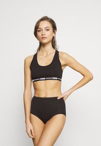 Zwarte sportbeha met een witte PUMA-logo band, gecombineerd met hoge zwarte onderbroek. Gladde stof, atletisch ontwerp, minimale details.