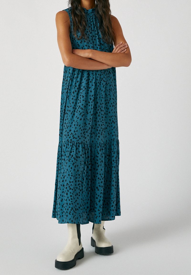 Femme portant une robe midi sans manches bleu sarcelle à pois noirs et des bottines blanches à semelles épaisses noires, debout les bras croisés.