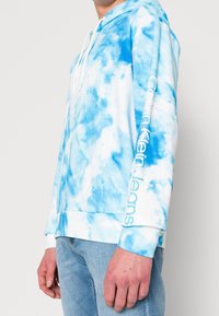 Felpa con motivo tie-dye blu e accenti bianchi, dotata di tasca frontale, cappuccio con cordini e logo stampato sulla manica.