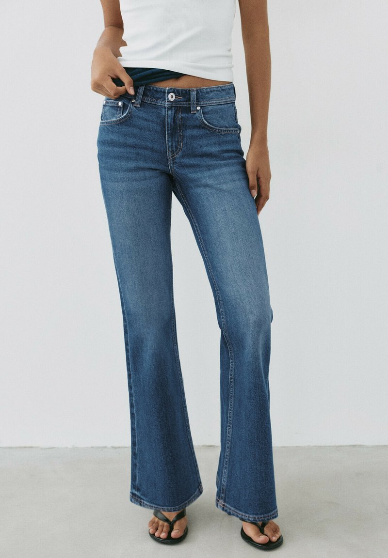 Personne portant un jean flare bleu taille haute, un haut blanc sans manches et des tongs noires, debout devant un fond clair uni.