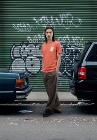 Orange bomulds T-shirt med grafisk print, løse brune bukser med nålestriber og sorte sko. Baggrunden har grøn graffiti og parkerede biler.