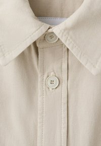 Col de chemise beige avec deux boutons blancs attachés, montrant une fine texture de tissu diagonale et des coutures cousues.