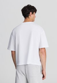 Biała koszulka z krótkim rękawem, z okrągłym dekoltem i luźnym krojem, prezentowana z tyłu, charakteryzująca się gładką fakturą i minimalistycznym wzornictwem.