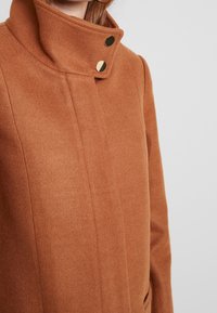 Bruine wolmix jas met een hoge kraag, twee goudkleurige drukknoopjes en een gestructureerde pasvorm met strakke lijnen.