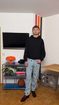 Giovane uomo con felpa nera con cappuccio e jeans ricamati si trova in una stanza con una TV, una lampada arancione, un pallone da basket, libri e una valigia argentata decorata con adesivi.