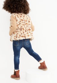 Faux fur-väst med leopardmönster, beige långärmade tröja, blå skinny jeans och bruna ankelskor med vågformad kant, på ett barn.