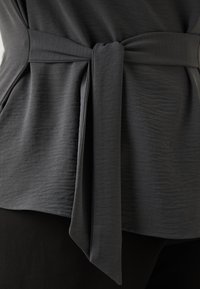 Dark gray top with a tied waist detail, featuring a textured fabric and smooth edges, layered over black pants.
