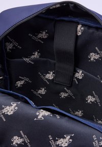 Inredningen av en marinblå ryggsäck med slätt tyg som har ett upprepat mönster med silverlogotypen för U.S. Polo Assn. och ett centralt svart webbinghandtag.