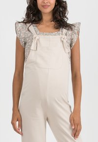 Beige overalls in soft fabric with adjustable straps and front pocket, paired with a floral short-sleeve top featuring ruffled sleeves.