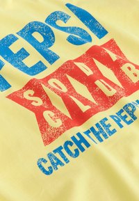 Geel katoenen t-shirt met blauwe en rode verweerde grafische tekst: "PEPSI SODA CLUB VANG DE PEPSI" in een vet, blokkerig ontwerp.
