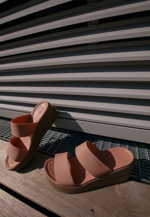 Keilabsätze mit zwei breiten Obergurten aus weichem Pfirsichmaterial, mit einer strukturierten Sohle, montiert auf einer hellbraunen Basis, vor einem laminierten Hintergrund.