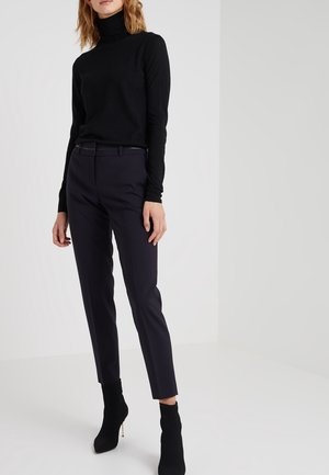 Femme portant un pull à col roulé noir, un pantalon sombre sur mesure et des bottines noires à talons, debout contre un mur blanc uni.