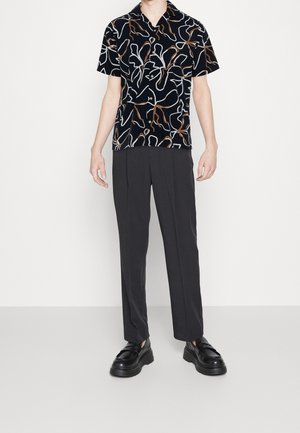 Maglietta nera a maniche corte con motivi astratti bianchi e marroni, abbinata a pantaloni scuri e scarpe nere chunky in un ambiente minimale.