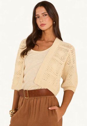 Vrouw met lang bruin haar draagt een crèmekleurig gehaakt vest over een beige top, bruine high-waist broek met brede riem en gouden armbanden.