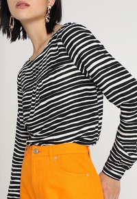 Černobílé pruhované dlouhé tričko s kulatým výstřihem, spárované se zářivě oranžovými kalhotami s vysokým pasem, které mají klasický střih a texturu.