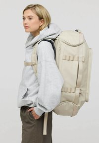 Zaino beige con molteplici scomparti, spallacci imbottiti e tasche laterali. Realizzato con materiale resistente, presenta una base arrotondata e spallacci regolabili.