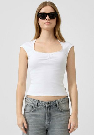 Vrouw met zwarte vierkante zonnebril, wit nauwsluitend topje met korte mouwen en vierkante halslijn, en hoogwatervervaagde grijze jeans.