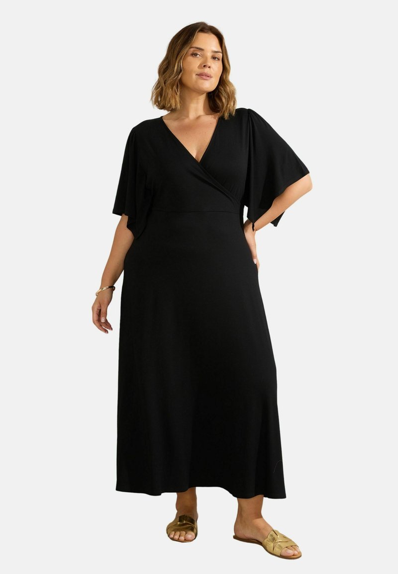 Robe noire enveloppante avec des manches courtes et légères. Tissu lisse, longueur à la cheville, décolleté en V modeste. Le mannequin porte des sandales à glissière dorées.