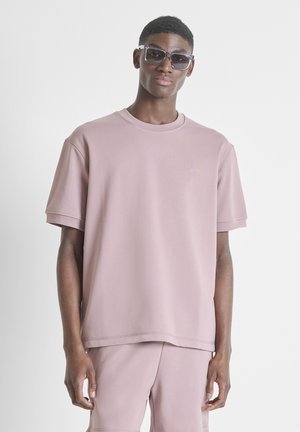 Giovane uomo che indossa una t-shirt oversize color malva chiaro e pantaloncini coordinati, con occhiali da sole rettangolari scuri, in piedi su sfondo bianco neutro.