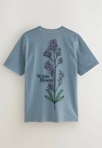 Mėlynas medvilninis marškinėliai su gėlių dizainu, kuriame yra rožinės gėlės ir žalios lapai nugaros pusėje. Tekstas sako "Wilde Bloem." Trumpi rankovės.