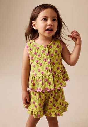 Mädchen mit braunen Haaren, das ein grünes ärmelloses Top und Shorts mit pinken Blumenmustern trägt, steht und hält ihr Haar.
