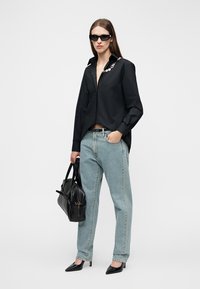 Femme portant une chemise noire avec un détail chaîne, un jean bleu clair, des talons noirs, des lunettes de soleil, et tenant un sac à main en cuir noir.