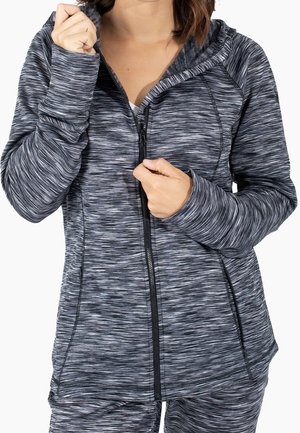 Femme portant un sweat à capuche zippé gris et noir rayé avec des trous pour les pouces, remontant la fermeture éclair sur un pantalon assorti.