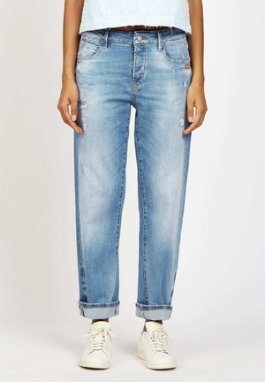 Person trägt hellblaue, umgeschlagene Jeans mit Used-Details und weiße Turnschuhe, steht vor einem schlichten Hintergrund.