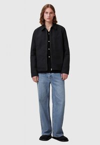 Sort letvægtsjakke med lynlås foran, to sidelommer og broderet detalje. Kombineret med løstsiddende blå jeans og sorte sko.