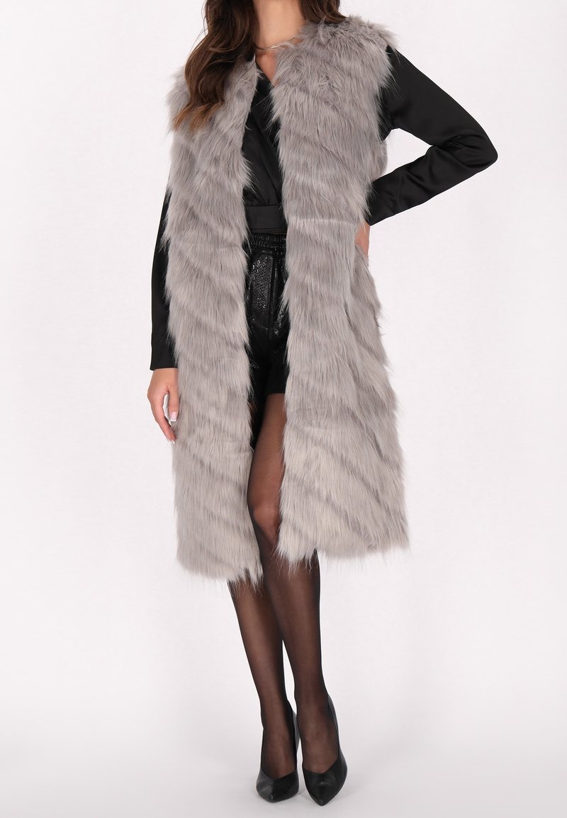 Graham Faux-Fur-Weste mit langem, offenem Design. Weicher, strukturierter Stoff mit flauschigem Aussehen, über einem schwarzen Outfit getragen und mit Absätzen kombiniert.