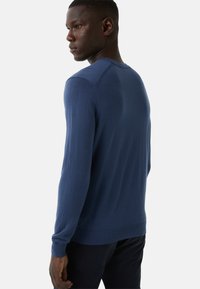 Mężczyzna ubrany w dopasowany niebieski sweter z długim rękawem, lekko odwrócony, na jednolitym białym tle.