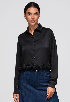 Femme aux cheveux foncés tirés en arrière portant une chemise noire en satin à manches longues boutonnée et une jupe en denim bleu taille haute sur un fond uni.