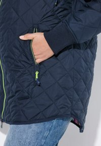 Giacca imbottita color marina con dettagli della cerniera verde e polsini a costine. Presenta tasche laterali e un orlo leggermente curvato.