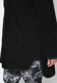 Primer plano de una chaqueta de forro polar texturizada negra con dobladillo redondeado sobre leggings negros con patrones florales blancos.
