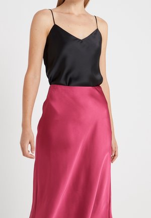 Femme portant un caraco en satin noir à fines bretelles, rentré dans une jupe taille haute en satin rose vif sur fond blanc.