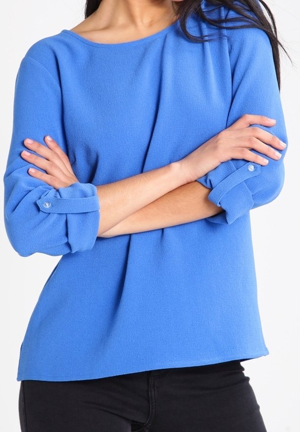 Femme bras croisés portant un haut bleu à manches longues avec manches retroussées et boutonnées et un pantalon noir.