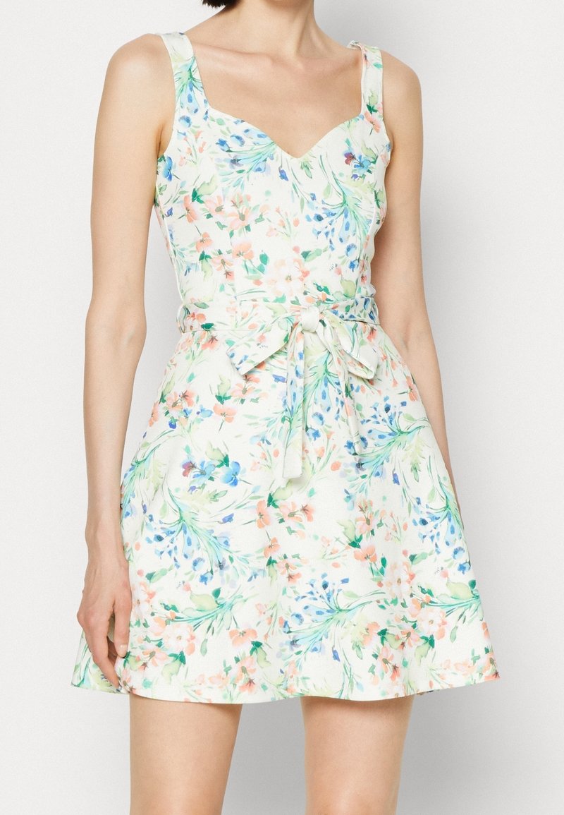 Robe blanche sans manches avec imprimé floral pastel, encolure en cœur et nœud à la taille, portée par une personne sur un fond uni.