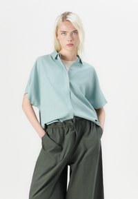 Světle modrá košile s krátkým rukávem a knoflíky, vyrobená z měkké tkaniny, spárovaná s volnými, tmavě zelenými kalhotami s elastickým pasem a bočními kapsami.