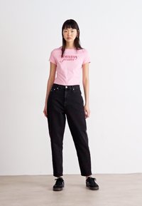 Rozā kokvilnas T-krekls ar "Tommy Jeans" logotipu, kombinēts ar augstās jostas melnām džinsu biksēm. Modelis ģērbjas melnās kedās. Neitrāla fona.