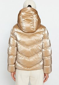 Giacca a vento metallizzata di colore beige con cuciture a zig-zag, un ampio cappuccio e una texture a coste. Presenta una vestibilità aderente e polsini elasticizzati.