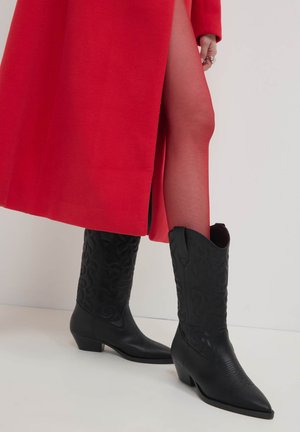 Stivali da cowboy neri con punta affusolata, decorazioni a rilievo e tacco basso, abbinati a un cappotto rosso e calze trasparenti.