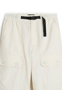 Pantaloni cargo beige con vita elastica, cintura nera con fibbia e due grandi tasche laterali, caratterizzati da una superficie liscia e dettagli cuciti.