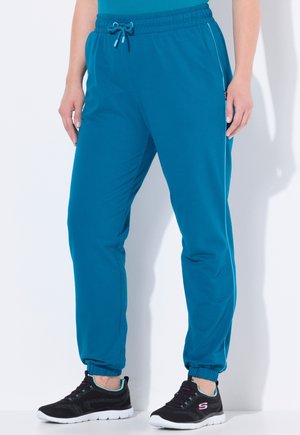 Teal Jogginghosen mit elastischem Bund und verstellbarem Kordelzug. Mit Seitentaschen und gerippten Knöcheln. Getragen mit schwarzen Sportschuhen.